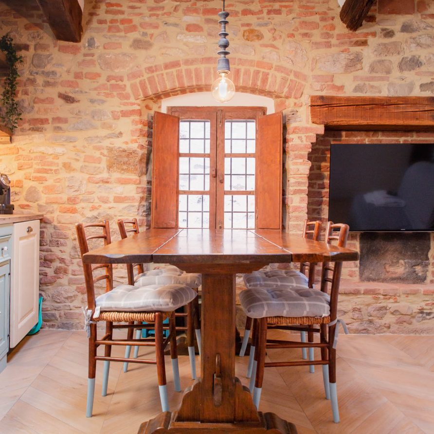 Interior of renovated holiday apartment showing stone walls exposed wooden beams and wooden dinning room table with four chairs next to bright window